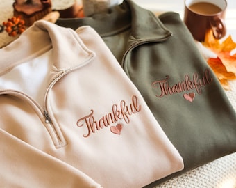 May include: Two quarter-zip sweatshirts in beige and olive green. The word "Thankful" is embroidered in a cursive font on the front of each sweatshirt, along with a small heart. The sweatshirts are folded and displayed on a soft, textured surface.