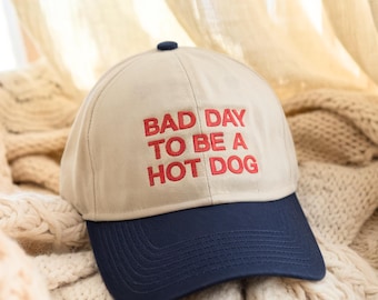 May include: A beige baseball cap with a navy blue brim. The front of the cap has red embroidered text that reads "Bad Day To Be A Hot Dog".