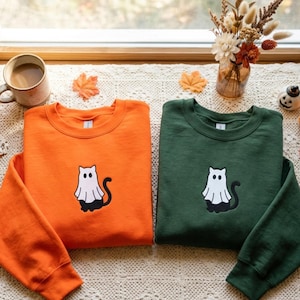 Peut inclure: Deux sweat-shirts à col rond, un orange vif et un vert forêt foncé, chacun arborant un motif brodé central représentant un chat noir vêtu d'un costume de fantôme blanc avec deux trous pour les yeux et une longue queue courbée.