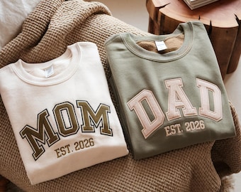 Peut inclure: Deux sweat-shirts col rond, un crème et un vert olive, sont présentés. Le sweat-shirt crème porte la broderie "MOM EST. 2026" en vert olive. Le sweat-shirt vert olive porte la broderie "DAD EST. 2026" en rose.