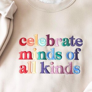 K&ouml;nnte beinhalten: Beiges Sweatshirt mit dem gestickten Text "celebrate minds of all kinds" in Regenbogenfarben. Der Text ist in einer fetten, serifenlosen Schriftart gehalten. Das Sweatshirt hat einen Rundhalsausschnitt und lange &Auml;rmel.