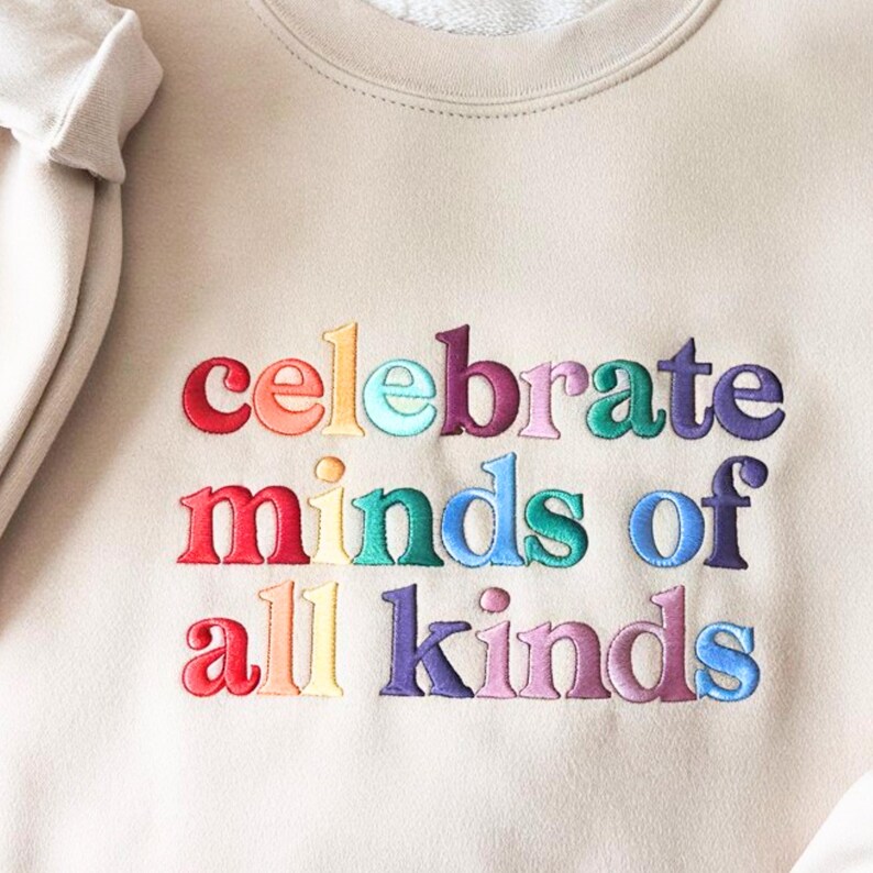 K&ouml;nnte beinhalten: Beiges Sweatshirt mit dem gestickten Text "celebrate minds of all kinds" in Regenbogenfarben. Der Text ist in einer fetten, serifenlosen Schriftart gehalten. Das Sweatshirt hat einen Rundhalsausschnitt und lange &Auml;rmel.