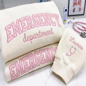 May include: Cream-colored sweatshirts with pink embroidered text. One sweatshirt says "EMERGENCY department" in a collegiate font. Another says "EMERGENCY". A third sweatshirt has the name "Lauren" and a stethoscope.
