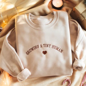 以下が含まれることがあります： ベージュのスウェットシャツ。茶色で「GROWING A TINY HUMAN」と刺繍され、小さな茶色のハートが添えられています。長袖のクルーネックトップスは柔らかい素材で作られています。