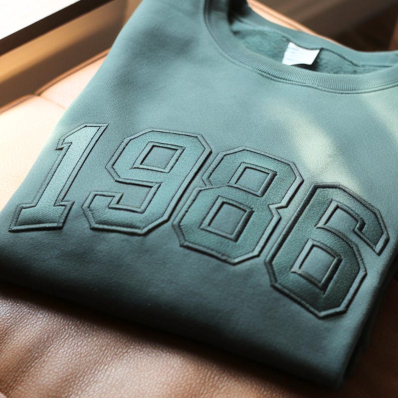 K&ouml;nnte beinhalten: Ein salbeigr&uuml;nes Sweatshirt mit Rundhalsausschnitt, das die Jahreszahl 1986 in gro&szlig;en, erhabenen, tonalen Stickbuchstaben auf der Brust zeigt. Das Kleidungsst&uuml;ck hat einen gerippten Kragen und ein weiches Fleecefutter, das am Halsausschnitt sichtbar ist.