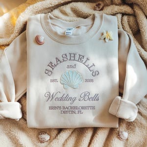 May include: A beige sweatshirt with the text "Seashells and Wedding Bells" embroidered on the front. The text is in a purple font with a seashell graphic in the center. The text "Erin's Bachelorette Destin, FL" is also embroidered below the main text.