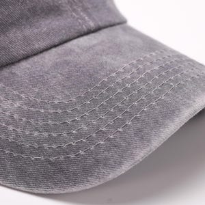 May include: Close-up of a gray baseball cap. The cap is made of a textured fabric and features a curved brim. White stitching details the brim and the seams of the cap, creating a subtle contrast against the gray material.