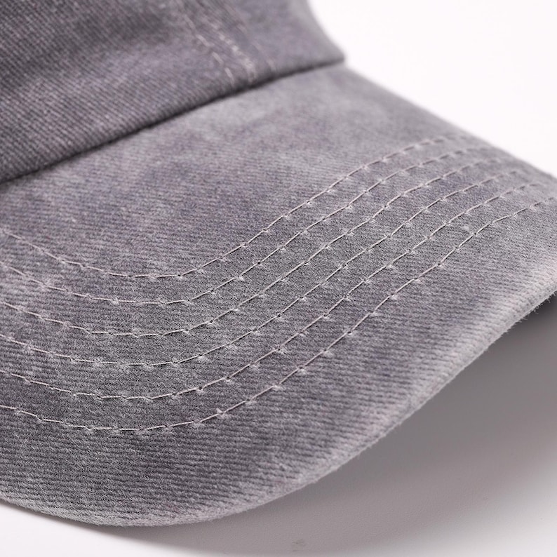 May include: Close-up of a gray baseball cap. The cap is made of a textured fabric and features a curved brim. White stitching details the brim and the seams of the cap, creating a subtle contrast against the gray material.