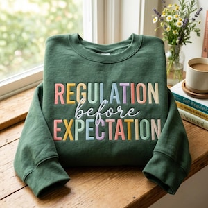 Könnte beinhalten: Ein dunkelgrüner Rundhals-Pullover mit gesticktem Text, der REGULATION before EXPECTATION lautet. Das Wort REGULATION ist in Blockbuchstaben, das Wort before in weißer Schreibschrift und EXPECTATION in Blockbuchstaben in Pastellfarben gehalten.