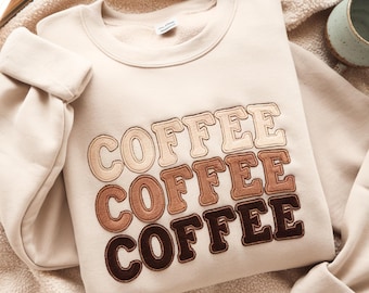 May include: Cream-colored sweatshirt with the word "COFFEE" embroidered in three rows of varying brown shades. The sweatshirt is folded, with the sleeves visible. The background is a soft, neutral color.