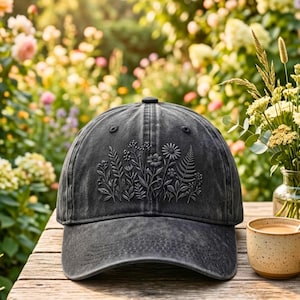 以下が含まれることがあります： 花柄のデザインが施された黒い野球帽。帽子には、花と葉の刺繍が施されています。帽子は、コーヒーカップと花瓶が背景にある木製のテーブルの上に置かれています。
