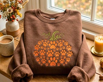 May include: A brown crewneck sweatshirt featuring an embroidered pumpkin design on the front. The pumpkin is composed of numerous small orange paw prints and topped with green embroidered vines and leaves, centered on the chest area.
