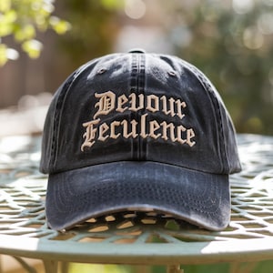 以下が含まれることがあります： 黒い野球帽。ベージュのゴシック体フォントで「Devour Feculence」の文字が刺繍されています。帽子は緑色の金属製のテーブルの上に置かれており、背景はぼやけています。