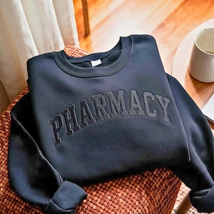 May include: A dark charcoal grey crewneck sweatshirt featuring the word PHARMACY embroidered in a matching dark thread across the chest in a bold, arched collegiate-style font. The garment has long sleeves and a ribbed collar neckline.