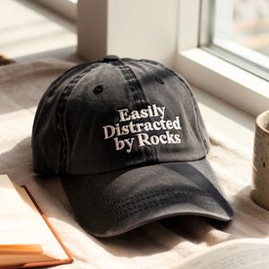 May include: A dark gray baseball cap with the embroidered text "Easily Distracted by Rocks" in white. The cap has a curved brim and a button on top. The hat is sitting on a beige surface.
