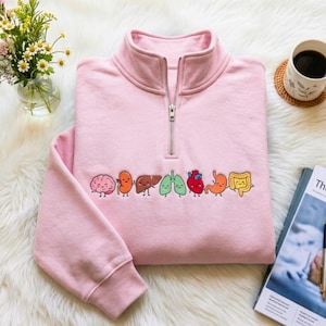 Puede incluir: Una sudadera rosa claro con cremallera de un cuarto que presenta una fila bordada de coloridas ilustraciones de órganos humanos sonrientes, incluyendo un cerebro, riñón, hígado, pulmones, corazón, estómago e intestinos.