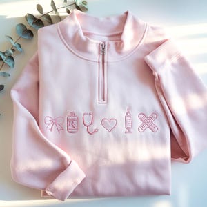 May include: A light pink quarter-zip sweatshirt with embroidered medical-themed icons. The design includes a bow, prescription bottle, stethoscope, heart, syringe, and bandages. The sweatshirt has a high collar and long sleeves.