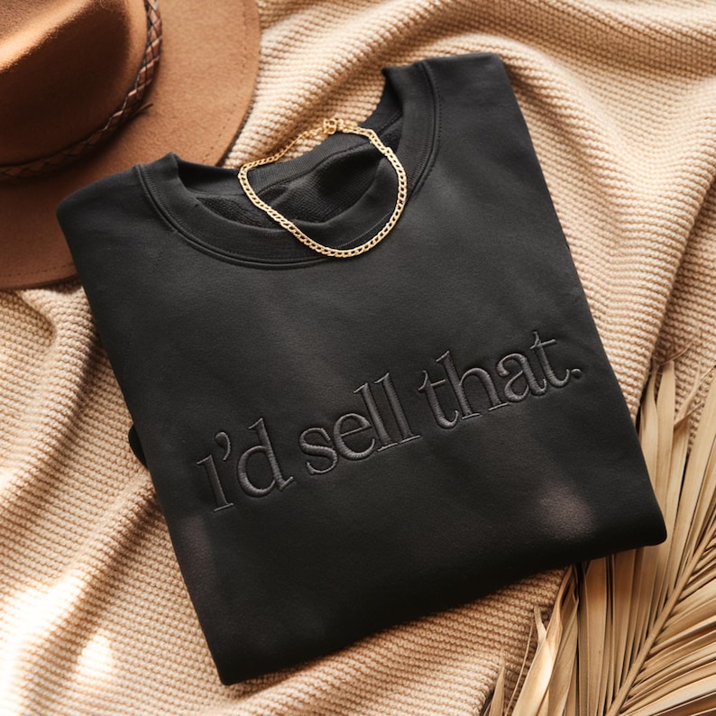 K&ouml;nnte beinhalten: Schwarzes Sweatshirt mit dem gestickten Satz "I'd sell that." Eine goldene Halskette liegt auf dem Sweatshirt. Das Sweatshirt ist auf einer beigefarbenen Strickdecke gefaltet, mit einem braunen Hut und einem Palmwedel im Hintergrund.