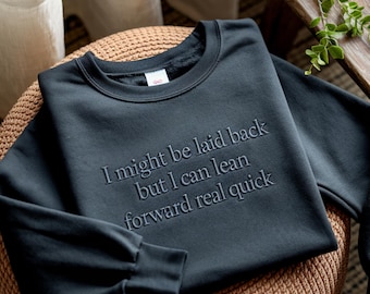 May include: A dark gray sweatshirt with the embroidered text: "I might be laid back but I can lean forward real quick." The crew neck sweatshirt is displayed on a woven surface.