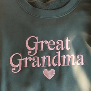 May include: A dark green sweatshirt with the embroidered words "Great Grandma" in pink script, with a pink heart below the text. The sweatshirt has a crew neck and a soft, fleecy interior.
