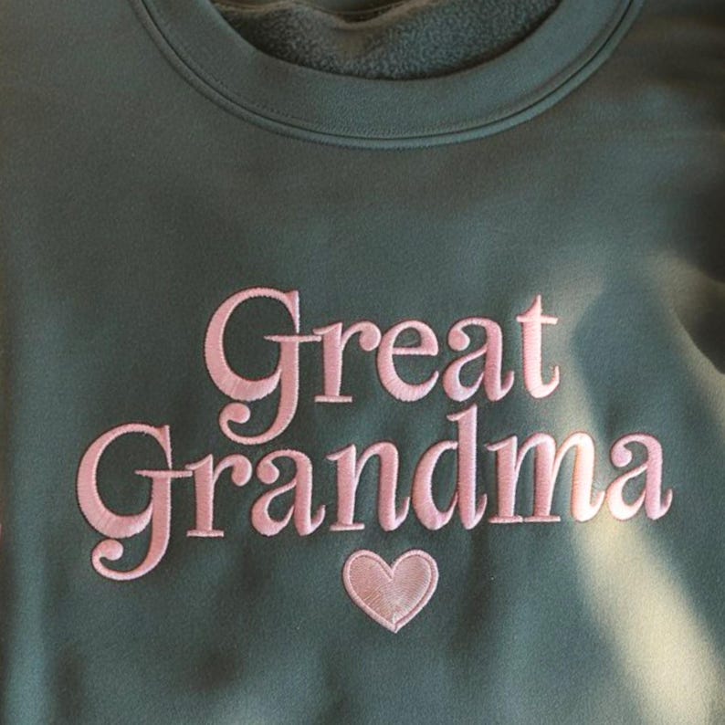 May include: A dark green sweatshirt with the embroidered words "Great Grandma" in pink script, with a pink heart below the text. The sweatshirt has a crew neck and a soft, fleecy interior.