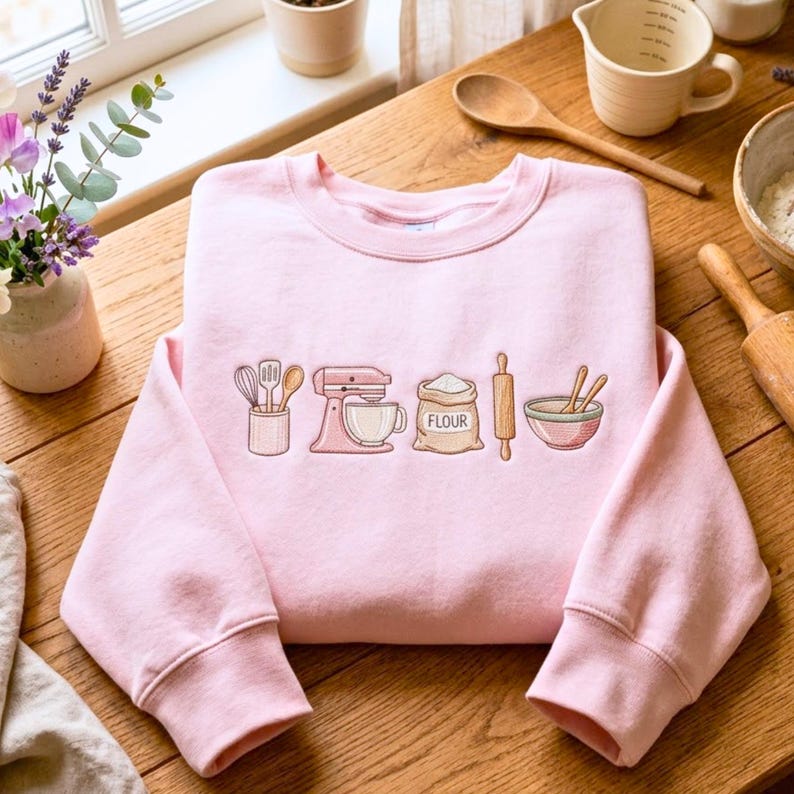 May include: A light pink crewneck sweatshirt featuring an embroidered design of kitchen baking tools including a utensil holder, a stand mixer, a bag labeled flour, a rolling pin, and a mixing bowl with spoons, centered on the chest area.