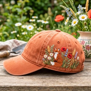 Könnte beinhalten: Eine rostorange Baseballkappe mit einer detaillierten Stickerei eines Fuchses inmitten von Wildblumen, darunter rote Mohnblumen, weiße Gänseblümchen und Weizenähren. Die Kappe verfügt über einen gebogenen Schirm und eine strukturierte Krone.
