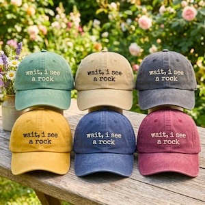 Könnte beinhalten: Sechs Baseballkappen in Grün, Beige, Anthrazit, Gelb, Blau und Kastanienbraun. Jede Kappe zeigt den gestickten Text "wait, i see a rock" in einer einfachen Schriftart. Die Mützen haben eine strukturierte Krone, einen gebogenen Schirm.