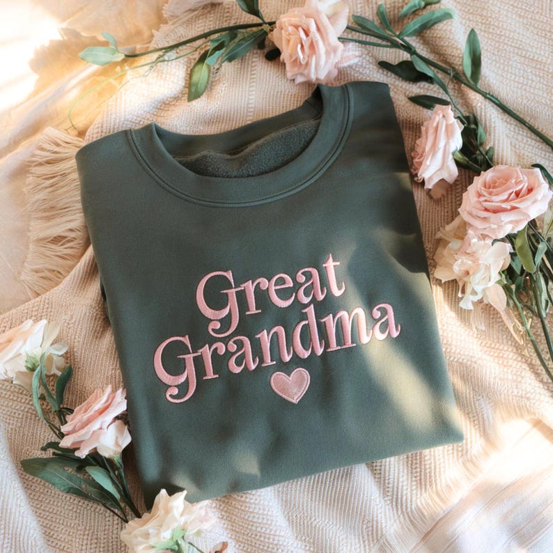 May include: An olive green sweatshirt with the embroidered words "Great Grandma" in pink script, along with a pink heart. The sweatshirt is surrounded by pink and white roses.