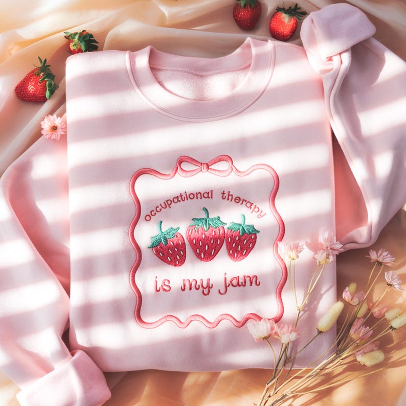 May include: A light pink sweatshirt with the embroidered phrase "occupational therapy is my jam" and three strawberries. The design is framed by a pink border with a bow at the top. The sweatshirt has long sleeves.