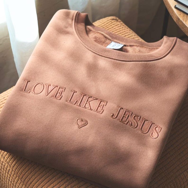 May include: A dusty rose-colored sweatshirt with the embroidered text "LOVE LIKE JESUS" and a heart symbol. The crew neck sweatshirt is folded and resting on a chair.