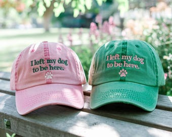 May include: Two baseball caps, one pink and one green, with the text "I left my dog to be here." embroidered on the front. Each cap has a paw print embroidered below the text.