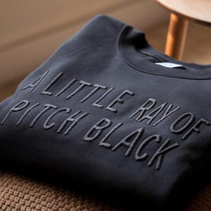 May include: A dark gray sweatshirt with the text "A little ray of pitch black" embroidered in black.