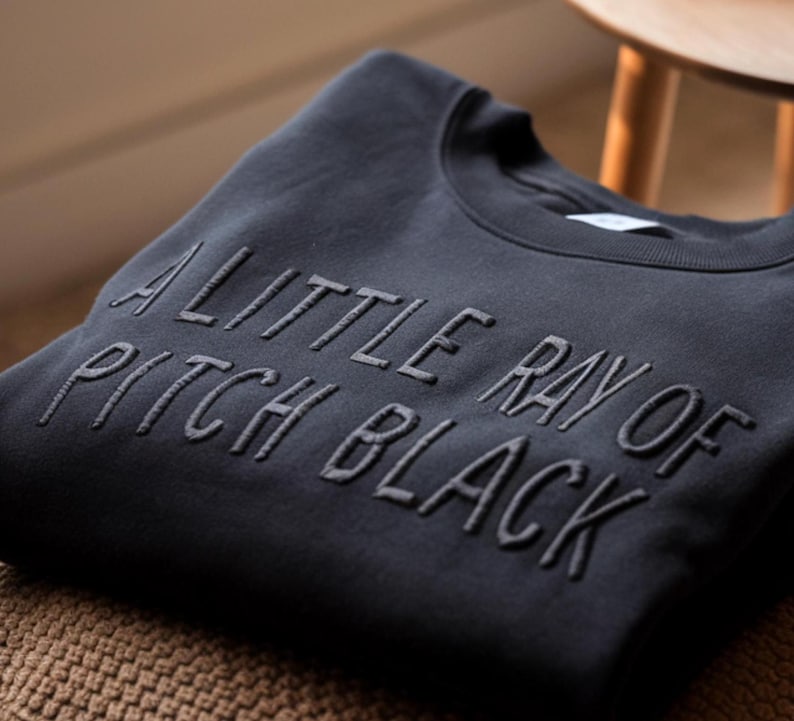 May include: A dark gray sweatshirt with the text "A little ray of pitch black" embroidered in black.