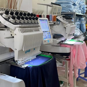 Puede incluir: Dos m&aacute;quinas de bordar blancas con m&uacute;ltiples agujas y pantallas digitales. Una m&aacute;quina tiene una tela azul marino en el bastidor, la otra una prenda rosa. Se ven carretes de hilo en el fondo.