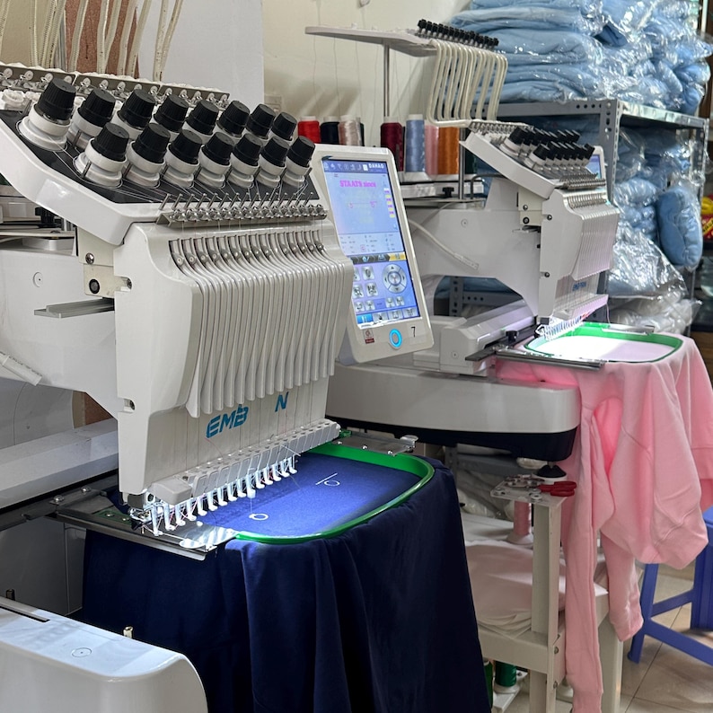 Puede incluir: Dos m&aacute;quinas de bordar blancas con m&uacute;ltiples agujas y pantallas digitales. Una m&aacute;quina tiene una tela azul marino en el bastidor, la otra una prenda rosa. Se ven carretes de hilo en el fondo.