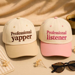 May include: Two baseball caps with text. One is cream-colored with a tan bill and the words "Professional yapper" embroidered in brown. The other is cream-colored with a pink bill and the words "Professional listener" embroidered in pink.