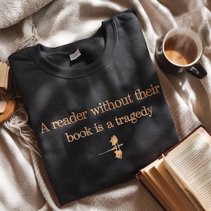 May include: A black sweatshirt with a gold embroidered quote: "A reader without their book is a tragedy". A small gold dragon design is below the quote.