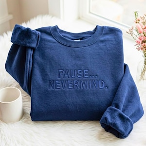 May include: A navy blue crewneck sweatshirt featuring the embroidered text PAUSE... NEVERMIND. in a matching dark blue thread. The garment has long sleeves with ribbed cuffs and a classic round neckline, showcasing a clean and simple design.