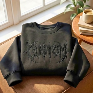 May include: A black crewneck sweatshirt featuring the word CUSTOM embroidered in a matching black, stylized, sharp-edged gothic font across the chest. The garment has long sleeves with ribbed cuffs and a ribbed neckline, presenting a monochromatic design.