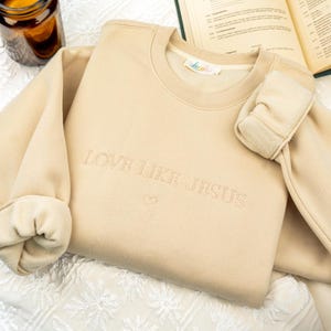 May include: A beige sweatshirt with the embroidered text "LOVE LIKE JESUS" and a small heart. The sleeves are cuffed, and the garment is resting on a white patterned surface. A small label is visible near the neckline.