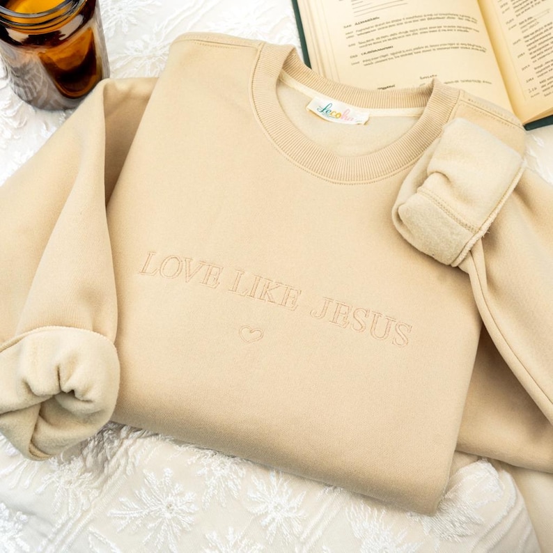 May include: A beige sweatshirt with the embroidered text "LOVE LIKE JESUS" and a small heart. The sleeves are cuffed, and the garment is resting on a white patterned surface. A small label is visible near the neckline.