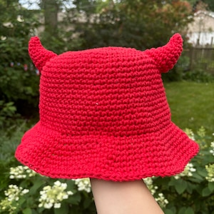 May include: A bright red, crocheted bucket hat with two curved horns on top. The hat has a wide brim and is made of a textured, woven material. The hat is held up against a backdrop of green foliage.