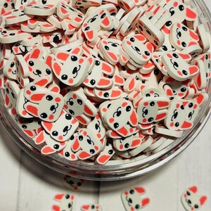 May include: A bowl filled with white and pink bunny face charms. The charms have black eyes and a black nose. The charms are perfect for adding a cute touch to crafts and projects.