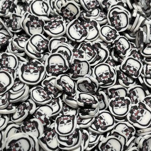 May include: A large quantity of white ceramic skull-shaped charms with black outlines and red details. The charms are scattered in a bowl.
