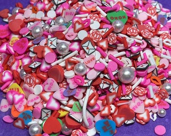 May include: A large pile of colorful Valentine's Day sprinkles, including hearts, letters, and other shapes. The sprinkles are in various shades of pink, red, white, and green.