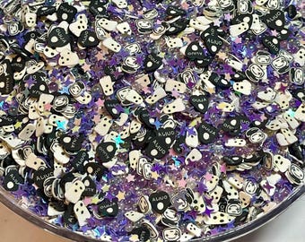May include: A close-up of a bowl filled with black and white plastic charms. The charms are shaped like ghosts, stars, and hearts. Some of the charms have the word "OUIJA" written on them. The bowl is filled with purple and iridescent glitter.