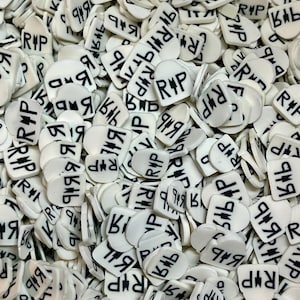 May include: A pile of white, tombstone-shaped beads with black text. The text includes the letters "RIP" and other symbols. The beads are scattered, creating a textured surface. The overall theme is related to memorial or remembrance.