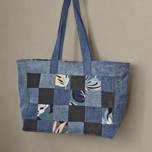 Peut inclure: Un sac fourre-tout en jean avec un motif patchwork. Le sac a une forme rectangulaire et des poignées robustes. Le patchwork incorpore des carrés de denim, de tissu gris foncé et de tissu à motifs avec des motifs de feuilles. Le sac est tenu par une main.
