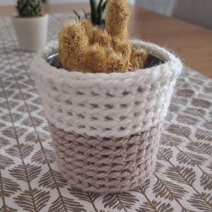 Peut inclure: Un petit cactus brun dans un pot en métal, niché dans un cache-pot tricoté à la main en deux tons. Le cache-pot est blanc en haut et marron clair en bas. Le cache-pot est posé sur une nappe à motifs.